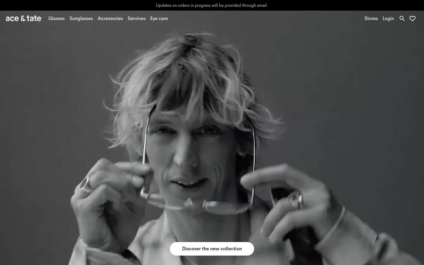 The desktop homepage features a large black and white video hero banner of a person adjusting glasses, with a white 'Discover the new collection' button centered at the bottom. A top navigation bar includes the Ace & Tate logo, product categories like Glasses and Sunglasses, and utility links for Stores, Login, Search, and Favorites. The layout is clean with a dark announcement bar at the very top.