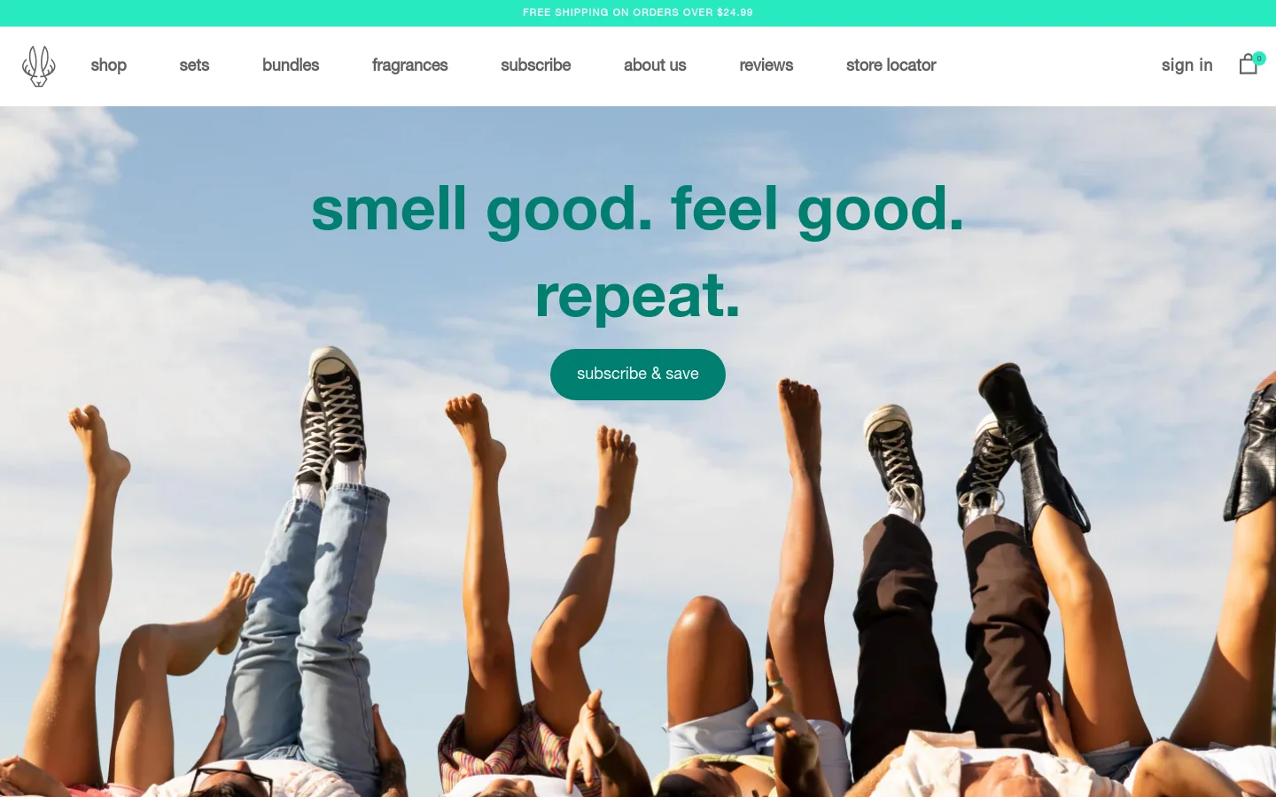 The desktop page features a large hero image of several people lying on grass with their legs raised toward a bright blue sky. Centered over the image is the headline 'smell good. feel good. repeat.' in teal text, followed by a teal 'subscribe & save' button. A white navigation bar at the top includes the brand logo and links for shop, sets, bundles, fragrances, subscribe, about us, reviews, and store locator.