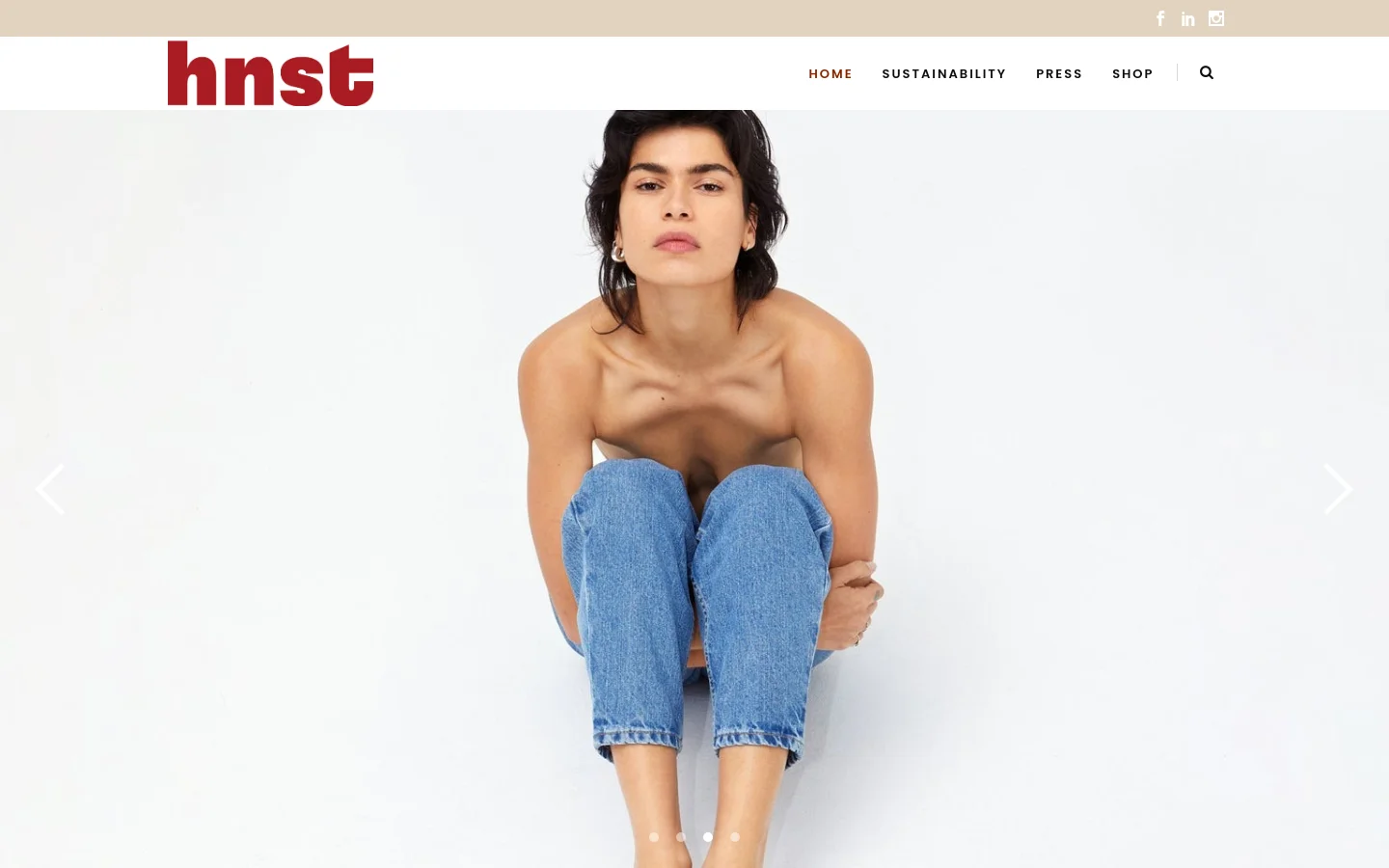 The desktop homepage features a minimalist design with a white background and a large hero image of a person sitting in blue jeans. A top navigation bar includes the HNST logo in dark red, links for Home, Sustainability, Press, and Shop, and social media icons. Navigation arrows on the sides of the hero image indicate a slider functionality.