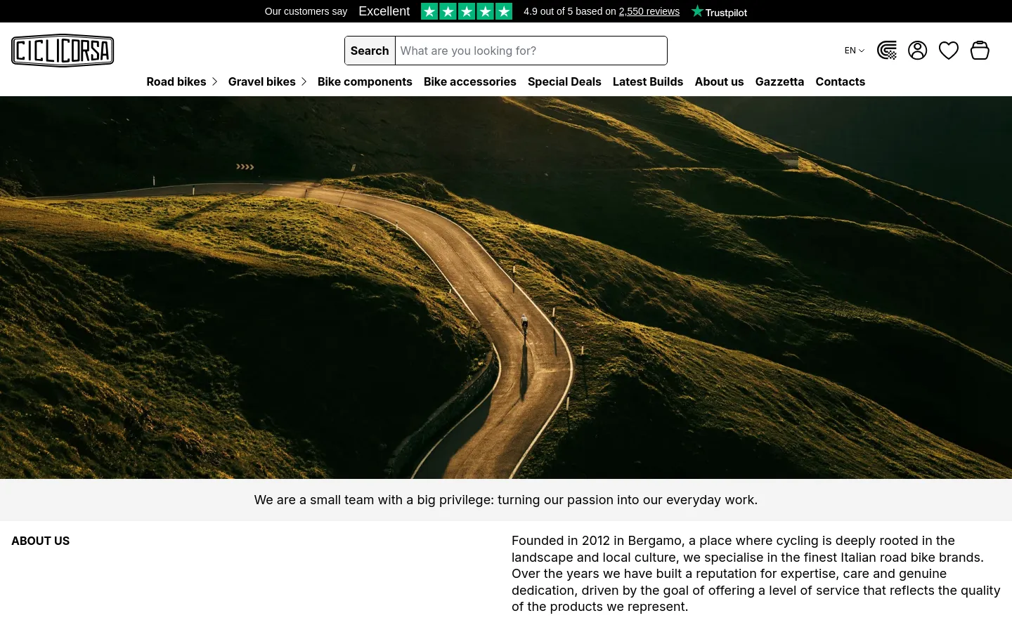 The desktop About Us page features a wide hero image of a winding mountain road at sunset with a lone cyclist. Below the image, a light gray banner contains a mission statement, followed by a two-column layout with the 'About Us' heading on the left and descriptive text on the right. The navigation menu is at the top, including a search bar and links for road bikes, gravel bikes, and components.