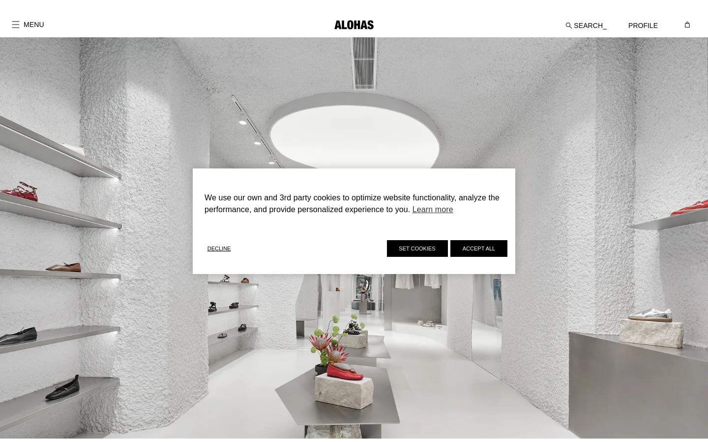 The desktop page features a minimalist, high-fashion aesthetic with a large hero image of a bright, white, textured interior boutique. A central white cookie consent overlay contains text and three buttons: 'DECLINE', 'SET COOKIES', and 'ACCEPT ALL'. The top navigation bar is clean, displaying the ALOHAS logo in the center with menu, search, profile, and shopping bag icons on the sides.