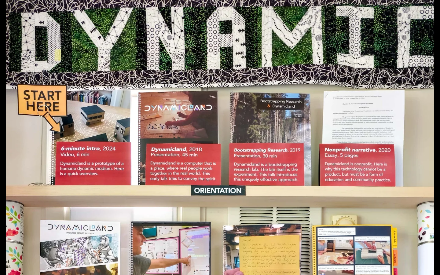 A desktop view of a digital bookshelf titled 'Dynamicland front shelf' against a light wood background. The top features a large, textured 'DYNAMICLAND' sign, followed by rows of various media items like videos, essays, and reports displayed as book covers or posters, and a bottom row of physical books. The layout is organized into sections like 'Orientation', 'Progress Reports', 'Dreams', and 'Everything'.
