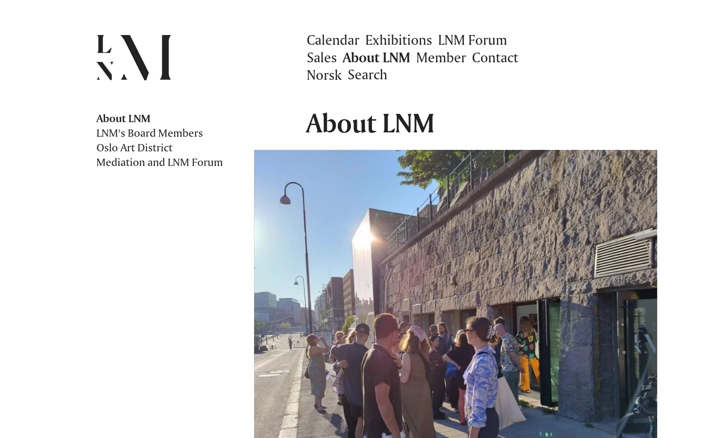 The desktop page features a minimalist design with a large LNM logo on the top left and a navigation menu on the top right. A large hero image shows a group of people standing outside a stone building in bright daylight under a clear sky. The main content area includes the headline 'About LNM' followed by text and a list of staff members.