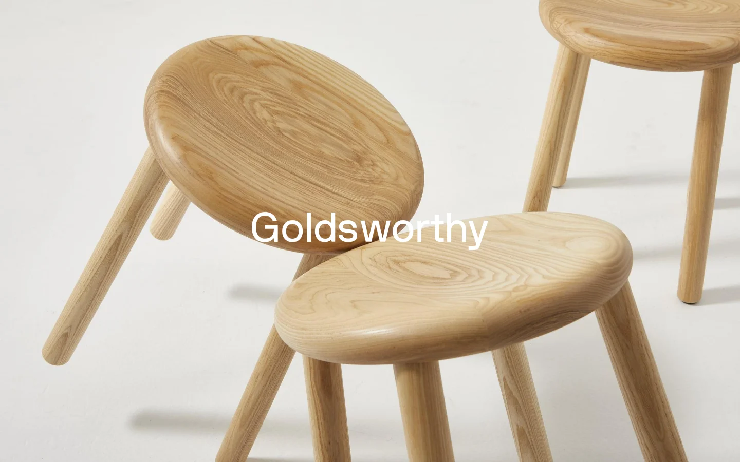 The desktop homepage features a minimalist design with a large hero image of light-colored wooden stools against a white background. The brand name 'Goldsworthy' is displayed in bold white text centered over the imagery, with a simple navigation menu at the top.