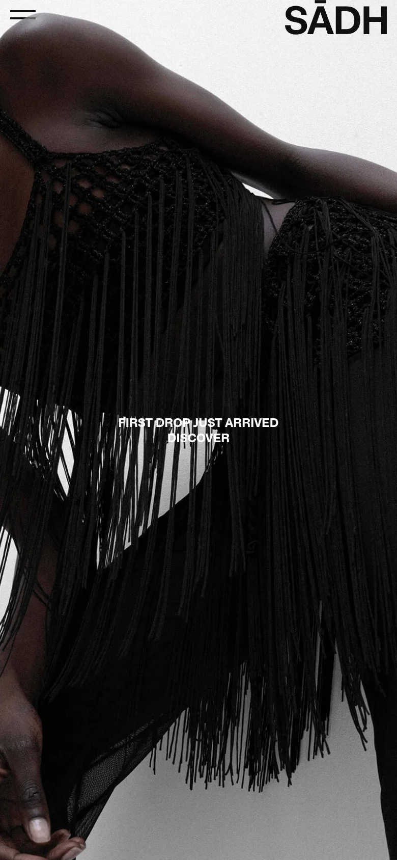 The mobile version presents a vertically oriented view of the same high-contrast black and white hero image, focusing closely on the textures of the black crochet fringe. The 'SĀDH' logo is positioned at the top right, and a hamburger menu icon appears at the top left. The 'FIRST DROP JUST ARRIVED' and 'DISCOVER' text is centered over the lower half of the image.