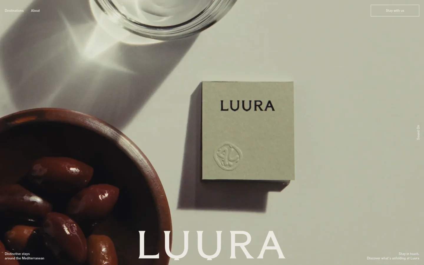 The desktop homepage features a minimalist design with a large, high-quality hero image of olives in a wooden bowl and a branded item on a neutral background. Large white serif typography displays the brand name 'LUURA' and the headline 'Distinctive stays around the Mediterranean.' A navigation bar at the top includes links for Destinations and About, alongside a 'Stay with us' call-to-action button.