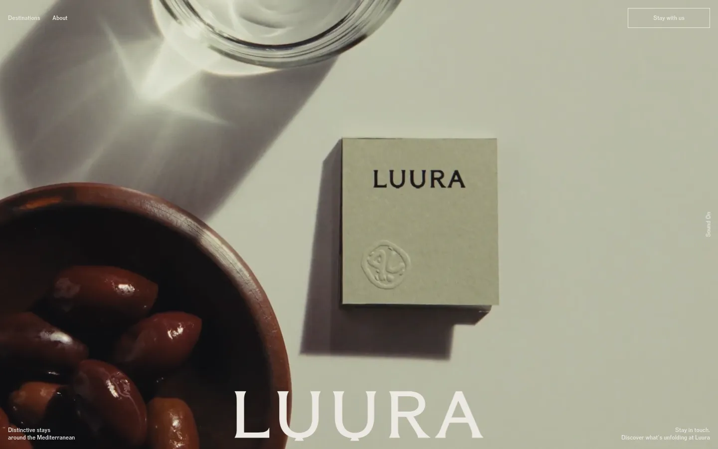 The desktop homepage features a minimalist design with a large, high-quality hero image of olives in a wooden bowl and a branded item on a neutral background. Large white serif typography displays the brand name 'LUURA' and the headline 'Distinctive stays around the Mediterranean.' A navigation bar at the top includes links for Destinations and About, alongside a 'Stay with us' call-to-action button.