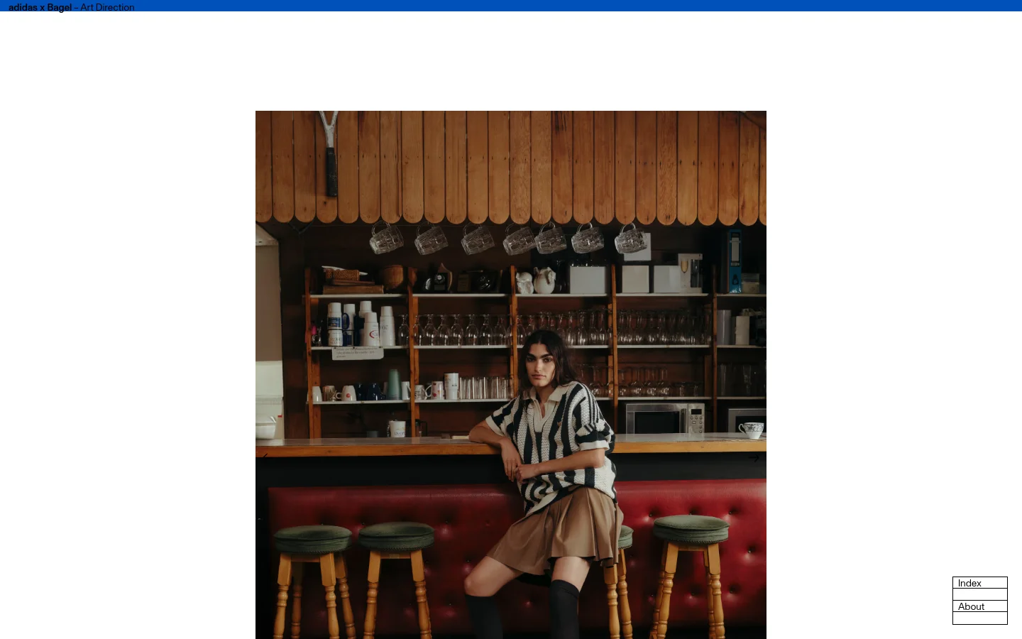 The desktop page features a minimalist layout with a large, full-width hero image of a person sitting at a wooden bar counter in a cafe setting. The headline 'adidas x Bagel – Art Direction' is positioned at the top left, and navigation buttons for 'Index' and 'About' are stacked in the bottom right corner against a white background.