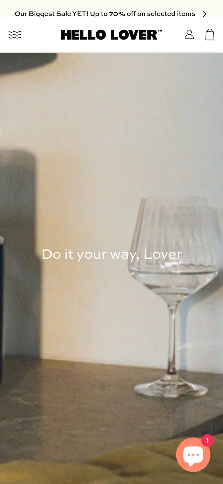 The mobile version presents a vertically stacked layout with a simplified header containing the Hello Lover logo, a menu icon, a user icon, and a shopping bag icon. The hero image remains prominent, featuring the 'Do it your way, Lover' text over a lifestyle photograph, and a floating chat bubble icon appears in the bottom right corner.