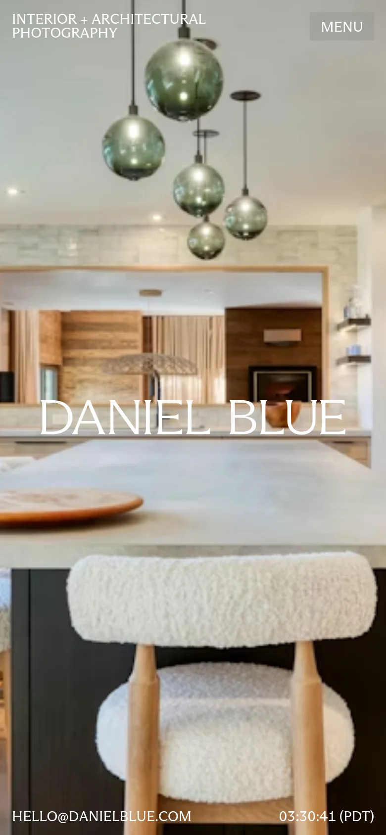 The mobile version presents a vertically oriented, zoomed-in view of the kitchen hero image, focusing on the green pendant lights and the textured white chair in the foreground. The branding 'DANIEL BLUE' is prominently centered, with a 'MENU' button in the top right and 'INTERIOR + ARCHITECTURAL PHOTOGRAPHY' in the top left. Contact information and the time are displayed in white text at the bottom of the screen.